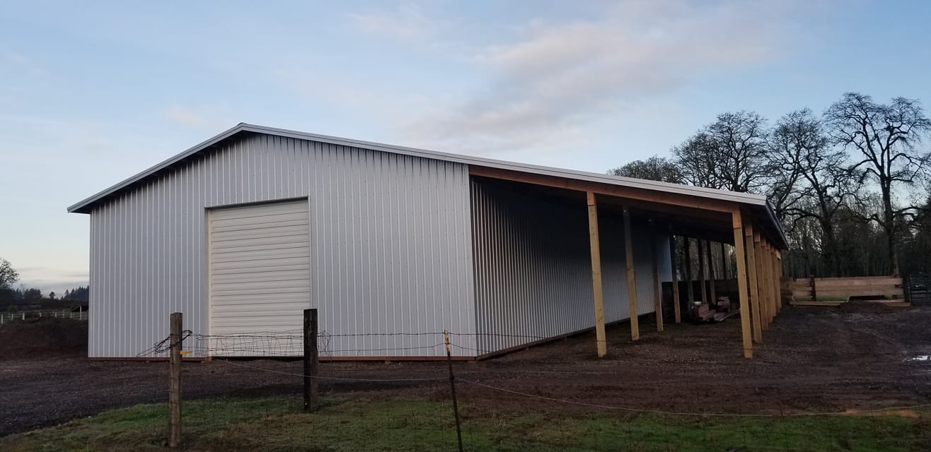 Picture of a large gray pole building with an overhead door and a lean-to on the side.